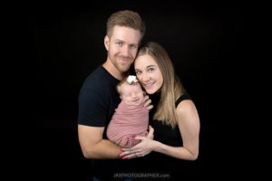 A man and woman smile while holding a newborn baby wrapped in a pink blanket, with a floral headband, against a black background—perfect for infant portraits in Jacksonville, Florida.
