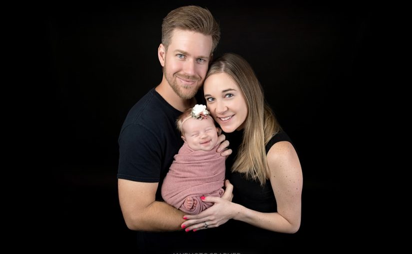 A man and woman smile while holding a newborn baby wrapped in a pink blanket, with a floral headband, against a black background—perfect for infant portraits in Jacksonville, Florida.