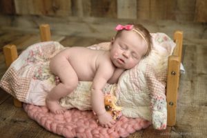 A sleeping baby with a pink bow lies on a small wooden bed with a quilt, holding a sprinkled donut. Perfect for Infant Portraits Jacksonville Florida, the baby's frosted face rests on a pink cushion over a wooden floor.