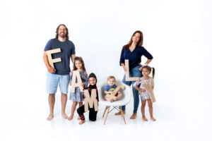 A smiling family of six poses on a white background, each holding large letters that spell “FAMILY.” Captured by a North Florida Family Photographer, the parents stand on either side, with four children in the middle, one sitting in a chair.