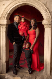 A family of four poses in front of a stone archway. Captured by a North Florida Family Photographer, the mother and daughter wear matching red dresses, while the father and son wear black suits with red accents. Everyone is smiling and holding hands.
