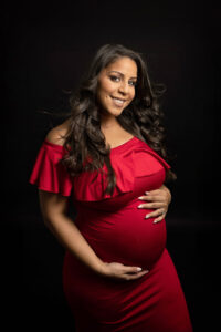 A smiling pregnant woman with long, wavy hair wears a red off-the-shoulder dress and cradles her baby bump with both hands against a dark background in a maternity photography studio Jacksonville.