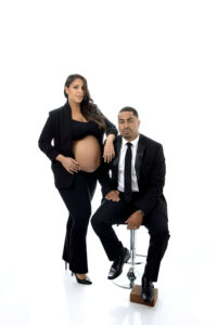 A pregnant woman in a black suit with an exposed belly stands beside a seated man in a black suit and tie, both elegantly posed against a plain white background by a skilled maternity photographer Jacksonville FL.