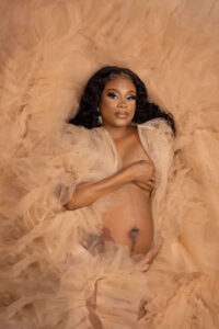A pregnant woman with long dark hair lies on soft, peach tulle fabric. She covers her chest with one hand and rests the other on her belly, gazing confidently at the camera in a maternity photography studio in Jacksonville.