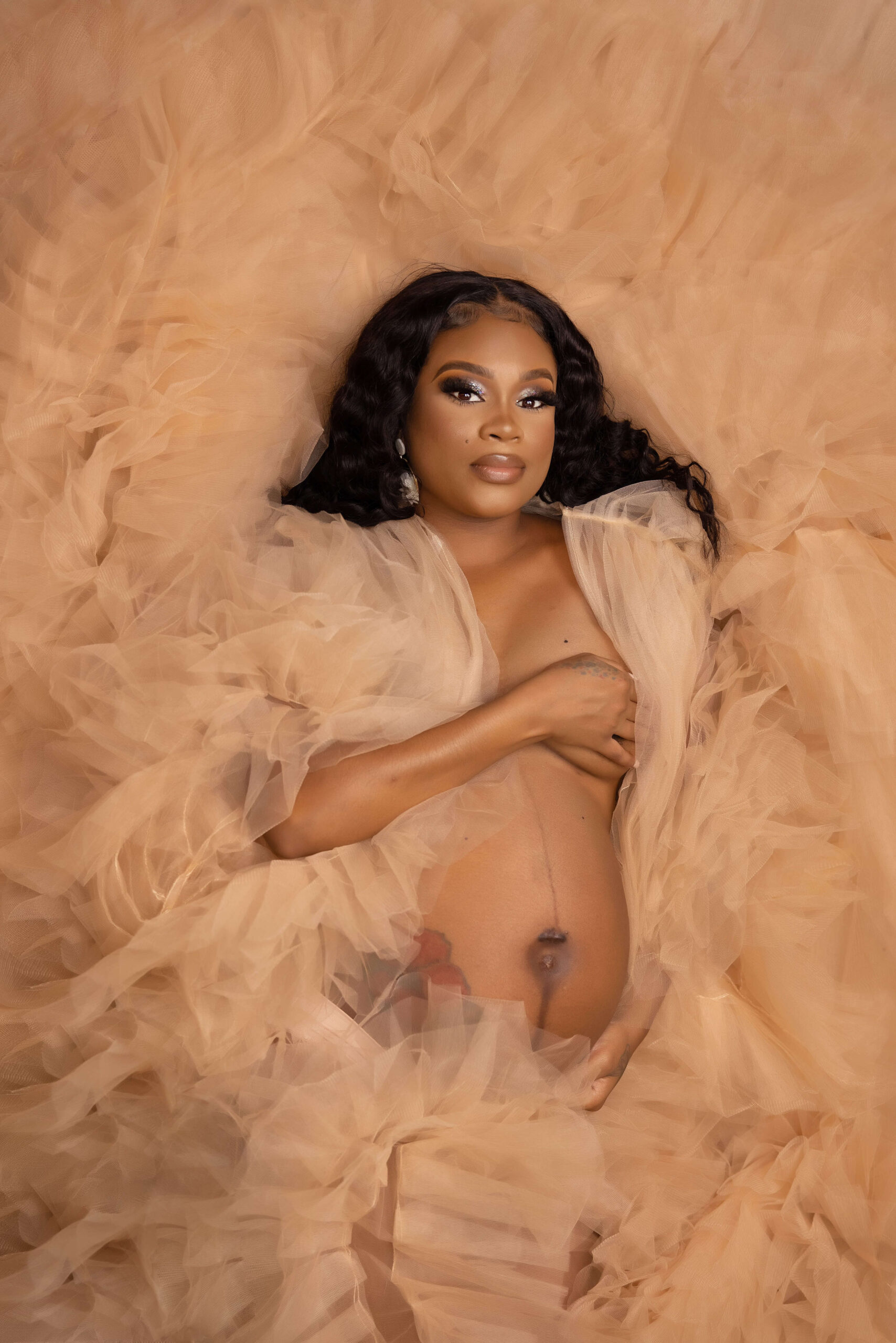 A pregnant woman with long dark hair lies on soft, peach tulle fabric. She covers her chest with one hand and rests the other on her belly, gazing confidently at the camera in a maternity photography studio in Jacksonville.