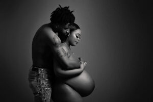 A shirtless man embraces a pregnant woman from behind, both standing against a dark background. The woman gently holds her bare belly, capturing an intimate and loving moment at a maternity photography studio in Jacksonville.