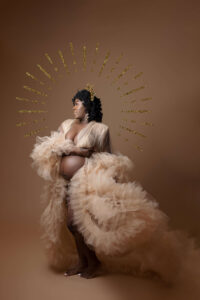 A pregnant woman in a sheer, ruffled gown stands against a brown background. She cradles her belly and wears a gold halo headpiece, styled by a maternity photographer Jacksonville FL, evoking a regal and ethereal appearance.