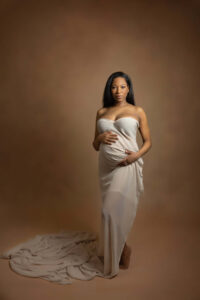 A pregnant woman with long dark hair stands barefoot against a brown backdrop, draped in white fabric. She holds her baby bump with both hands and looks calmly at the camera, beautifully captured by a Jacksonville baby photographer.
