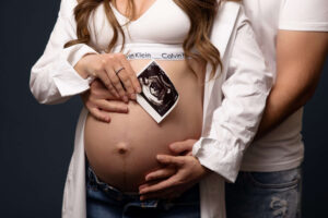 A pregnant woman holds an ultrasound photo over her bare belly, with a man's hands gently resting on her stomach. Both wear casual clothes, their faces unseen—captured intimately by a Jacksonville baby photographer.