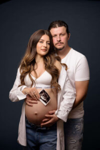 A pregnant woman in a white bra and open shirt holds an ultrasound photo on her bare belly, while a man stands behind her with his hands on her stomach. Both look at the camera—an intimate moment captured by the best maternity photographer Jacksonville.