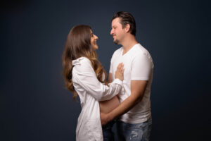 A pregnant woman and a man stand facing each other, smiling and holding hands. Captured by a maternity photographer Jacksonville FL, the woman wears a white shirt and the man wears a white t-shirt and jeans against a dark background.