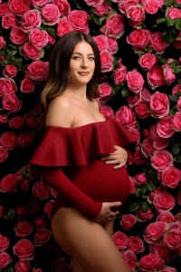 A pregnant woman wearing a red off-shoulder bodysuit poses in front of a wall covered with pink roses, holding her belly and smiling softly at the camera—captured beautifully by the best maternity photographer Jacksonville.