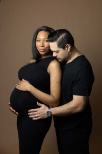 A pregnant woman in a black dress stands with a man beside her, his hands and head gently resting on her baby bump. Captured against a plain brown background, this North Florida maternity photography session highlights the best time for photos.