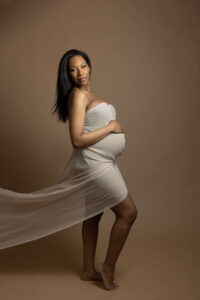 Pregnant woman in a strapless, sheer beige dress stands barefoot against a brown background, cradling her belly and looking at the camera, with fabric flowing behind her—captured by the best maternity photographer Jacksonville.