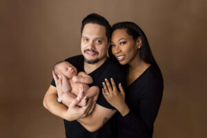 A man holds a sleeping newborn baby while standing beside a woman, all dressed in black against a plain brown background. The woman, showing her ring, rests her hand on his shoulder. Captured by a North Florida newborn photographer, all three are smiling.