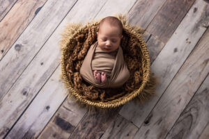 A newborn baby wrapped in a brown blanket is sleeping peacefully in a round woven basket on a rustic wooden floor, perfectly captured by a Jacksonville baby photographer.
