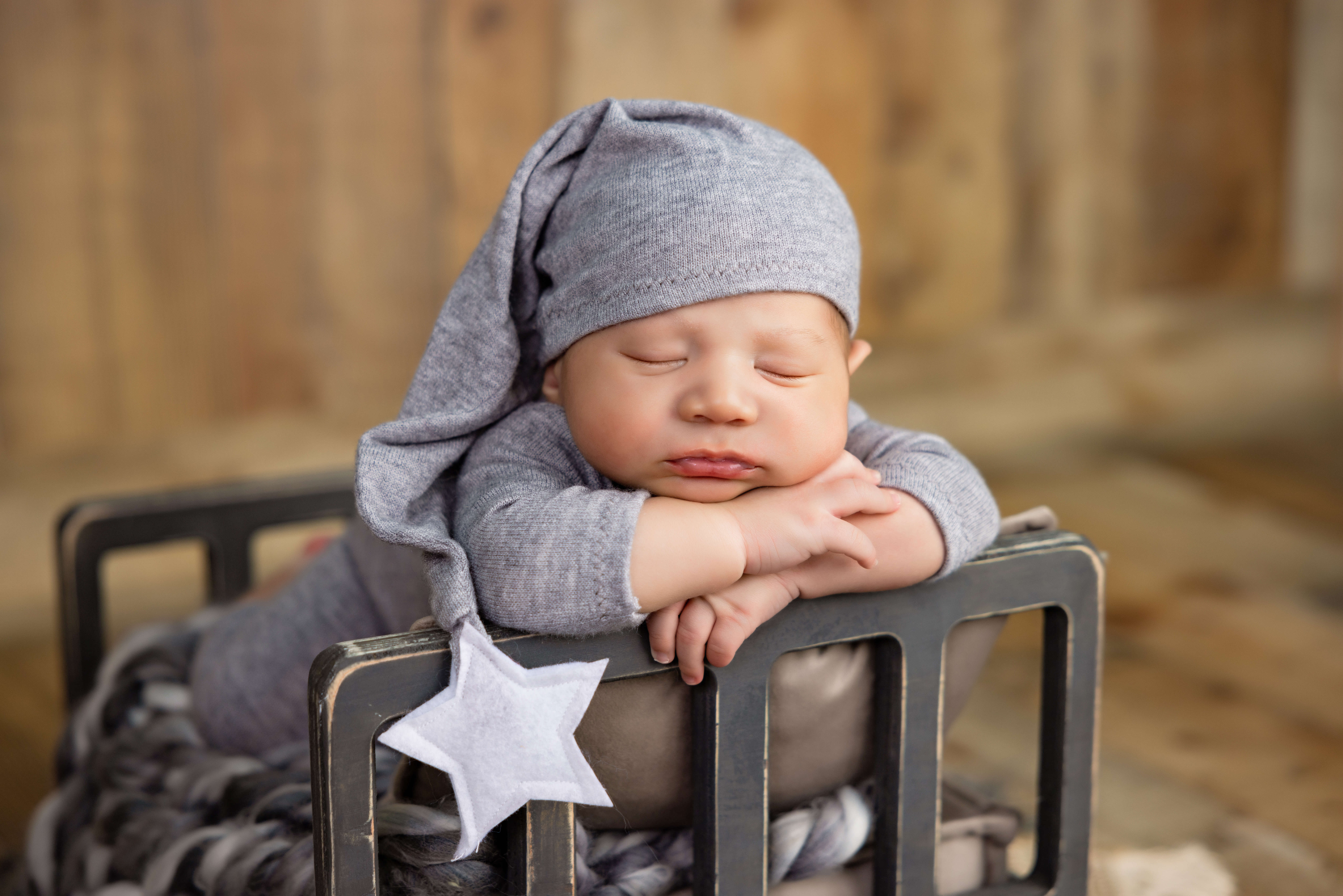 A sleeping baby wearing a grey outfit and a long hat with a star, resting on a small bed with a soft blanket, set against a warm, wooden background—captured by the best newborn photographer in Jacksonville, FL.