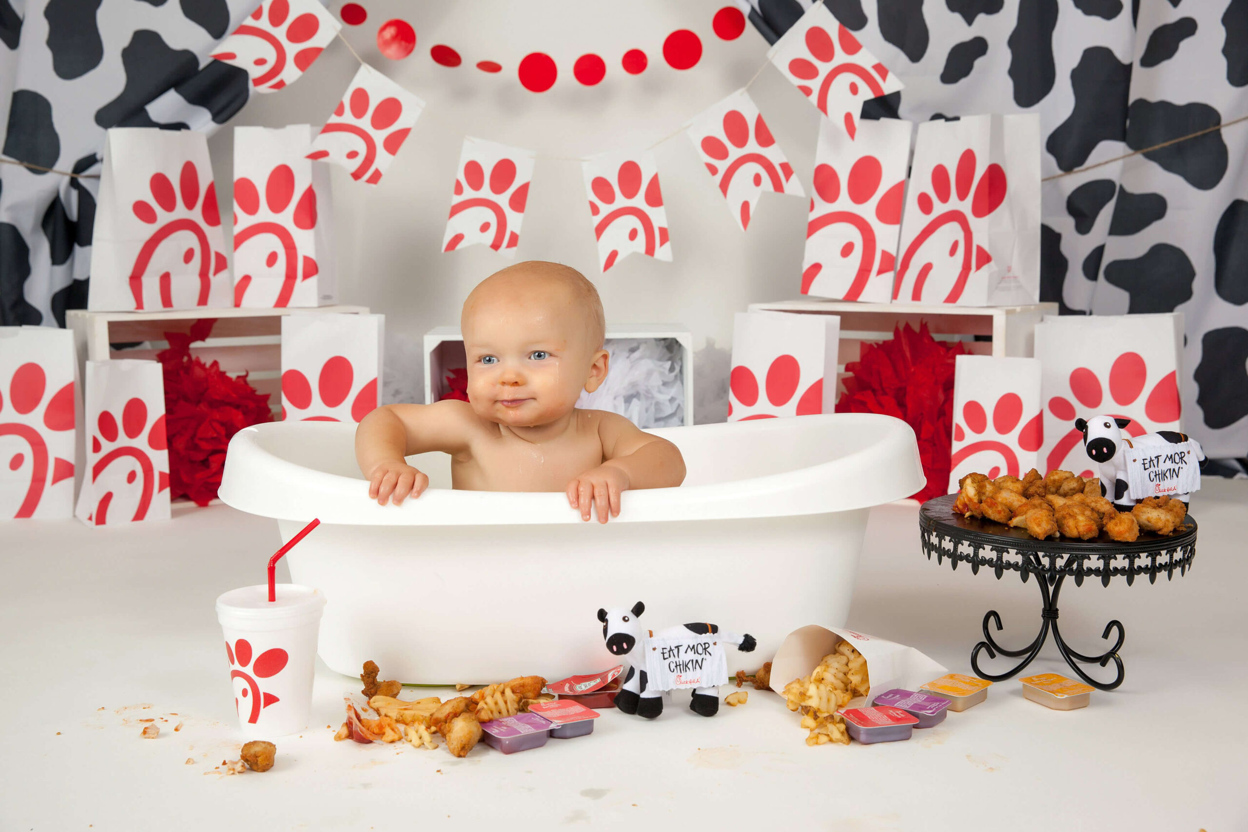 A baby sits in a white tub surrounded by Chick-fil-A food, cups, sauce packets, and branded bags—perfect for playful First Birthday Photos. Plush cows and nuggets are nearby amid a cow-pattern backdrop by Christy Whitehead Photography.