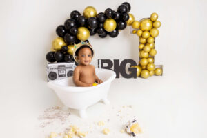 A smiling baby wearing a crown sits in a white clawfoot tub, surrounded by black and gold balloons forming the number one, with a "BIG 1" sign and toy boombox—perfect for a cake smash photographer Jacksonville FL to capture this fun milestone.