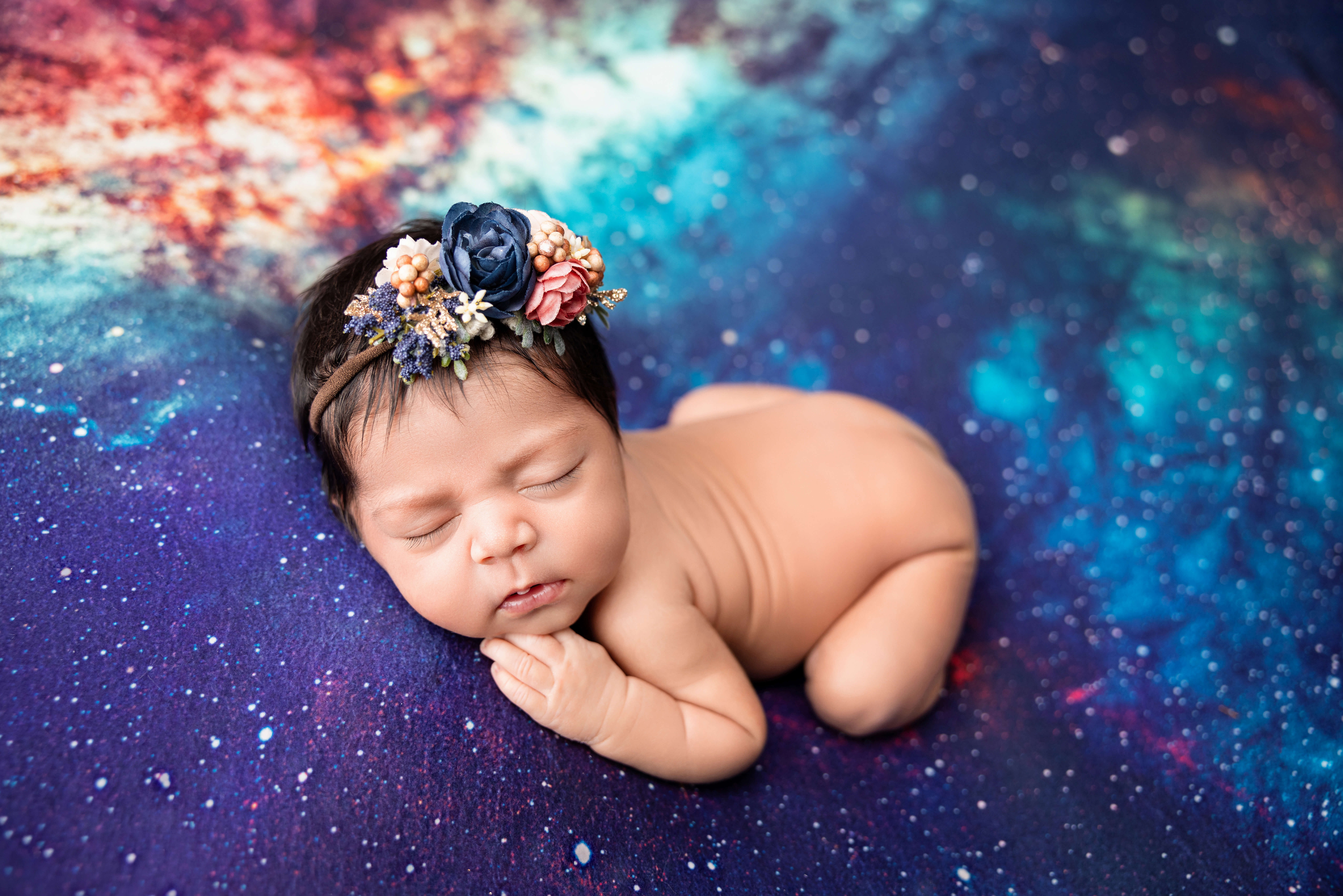Newborn baby sleeping on a colorful, galaxy-themed blanket, wearing a floral headband with blue, pink, and purple flowers. Captured by a newborn photographer Jacksonville FL—ask about the best time to book your session for dreamy photos like this!.