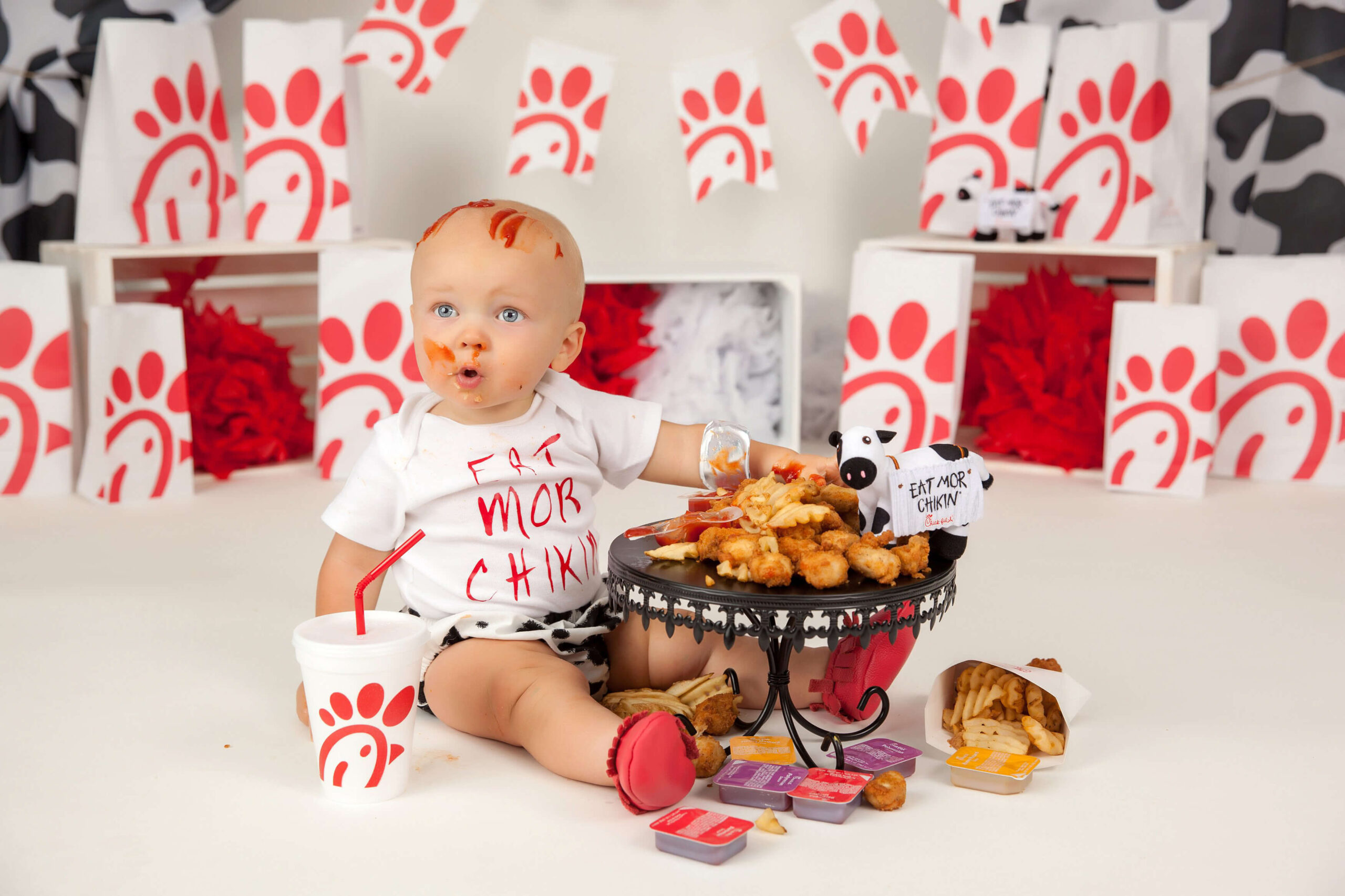 A baby wearing an "Eat Mor Chikin" shirt sits on the floor surrounded by Chick-fil-A bags and sauce, reaching for nuggets and fries—captured perfectly by a talented cake smash photographer Jacksonville FL.