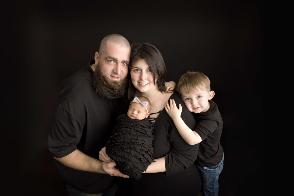 A family of four poses against a black background. The parents stand close together, holding their newborn wrapped in black as a young boy leans on his mother's shoulder. Everyone wears black clothing, capturing the warmth of Jacksonville Family Photography.