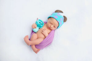 Newborn baby sleeping on a white blanket, wrapped in a purple cloth, wearing a blue and purple hat with horns, and holding a small blue stuffed toy—captured by your Jacksonville baby photographer.