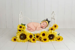 A newborn baby wearing a bee hat sleeps curled up on a white cushion in a small white crib, surrounded by bright yellow sunflowers—an adorable scene captured by a Jacksonville baby photographer.