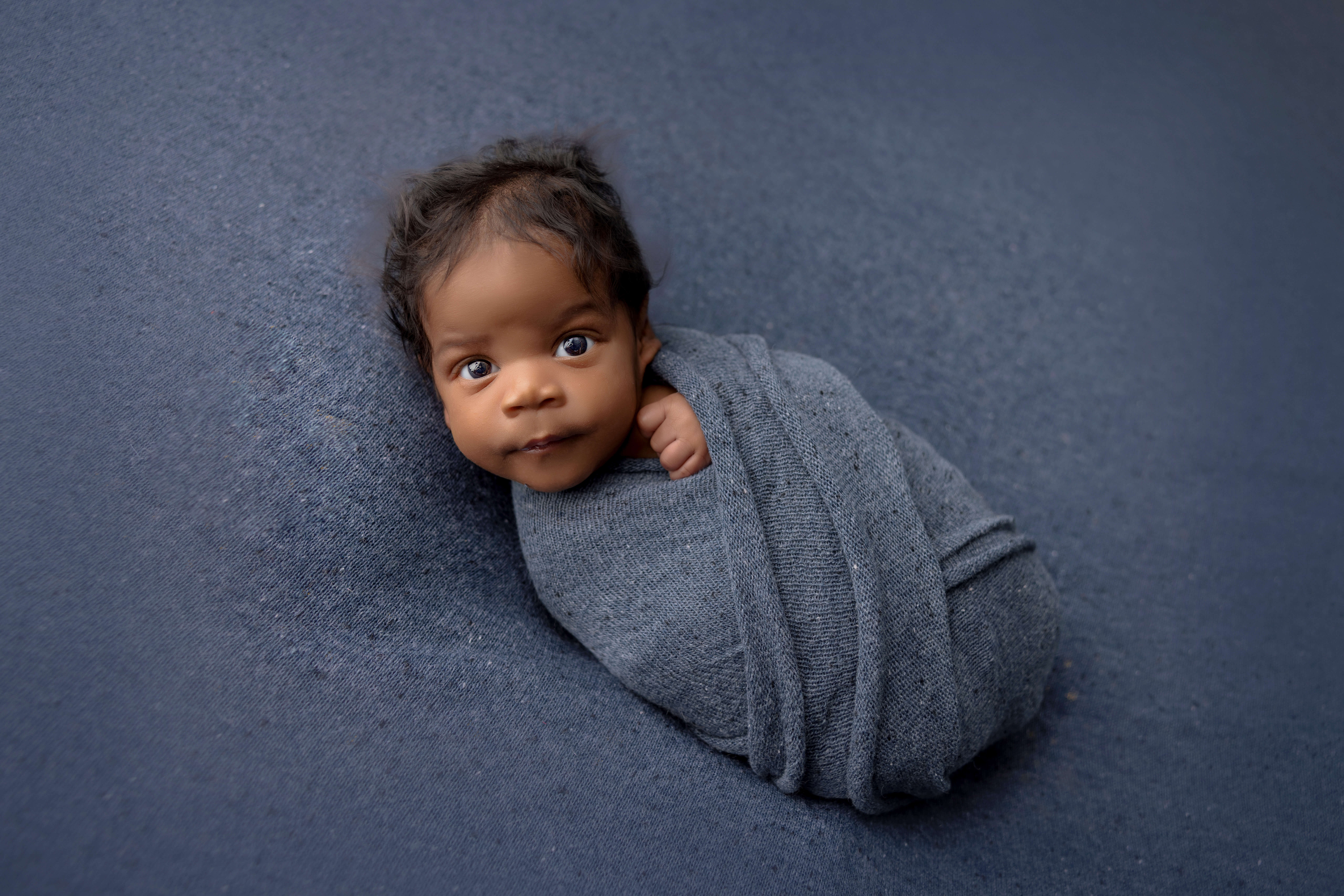 A baby wrapped snugly in a soft, gray blanket lies on a matching gray surface, looking up with wide, curious eyes—an adorable moment captured in classic Jacksonville newborn photography style.