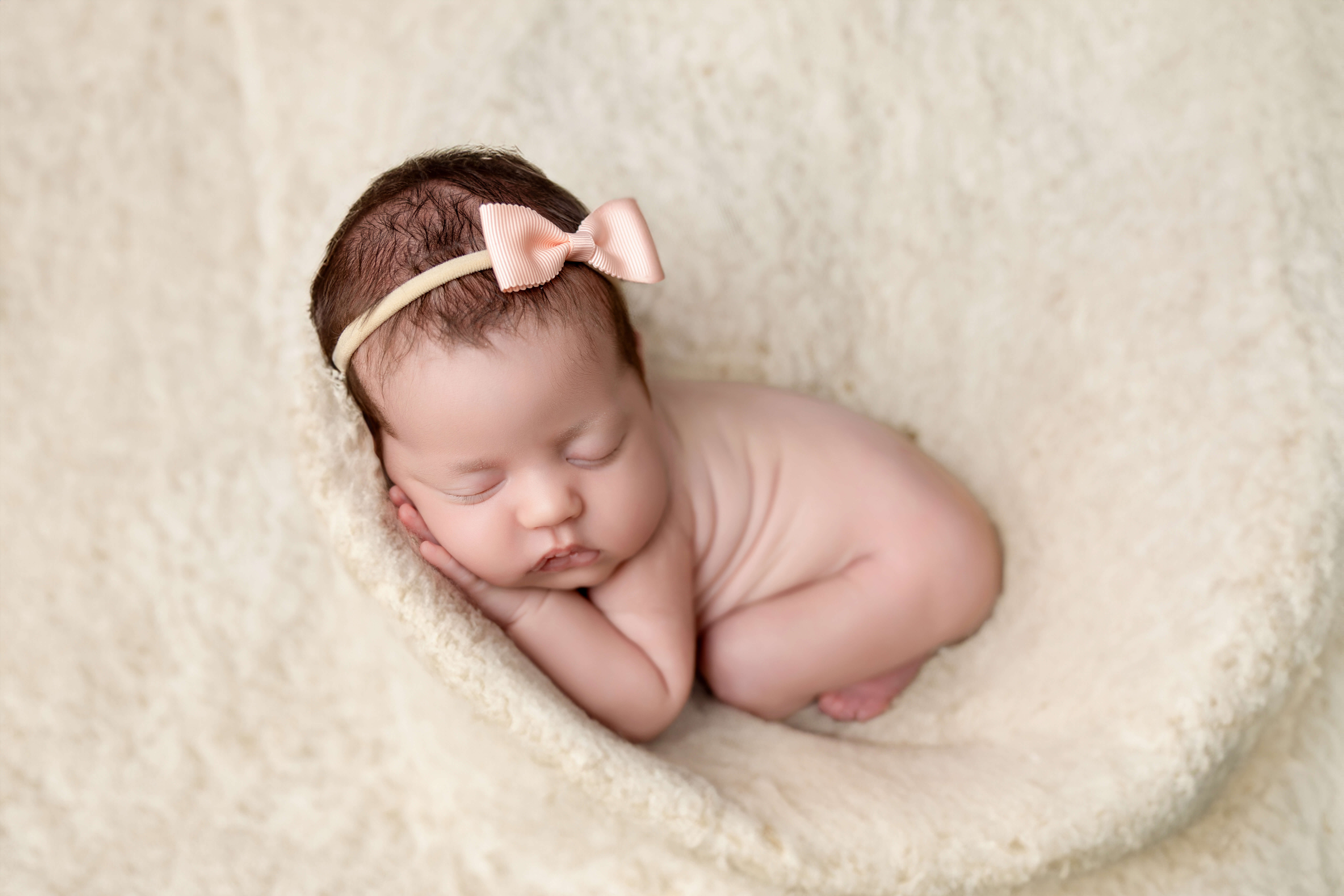A newborn baby with closed eyes sleeps curled up on a soft, cream-colored blanket, wearing a peach-colored bow headband—captured by a bump to baby photographer in Jacksonville, FL.