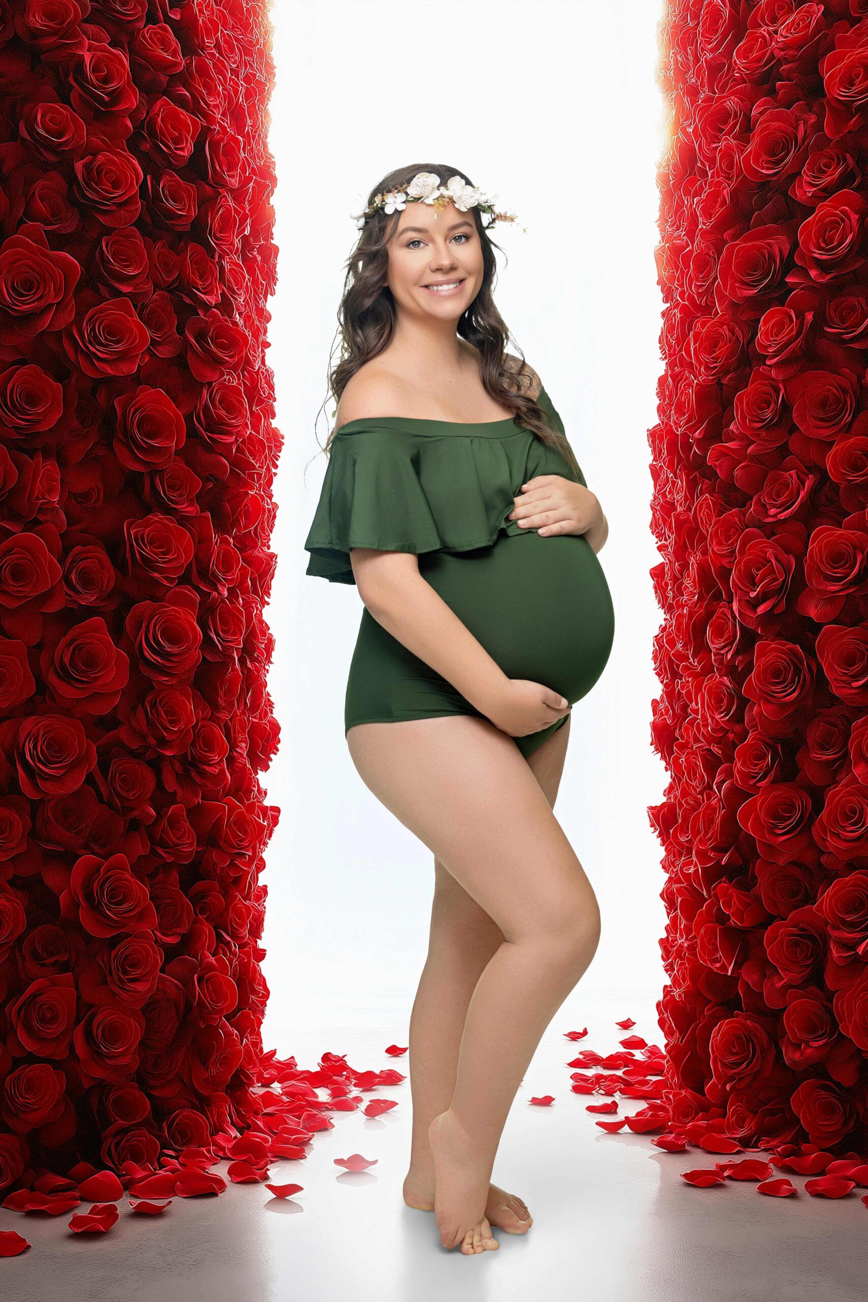A pregnant woman in a green off-shoulder bodysuit and flower crown stands barefoot between two tall walls of red roses, smiling and holding her belly. Red rose petals are scattered on the white floor in this maternity photography studio Jacksonville scene.