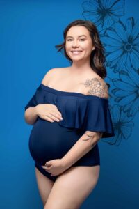 A smiling pregnant woman in a navy blue off-shoulder bodysuit poses against a blue background with floral illustrations, showcasing her tattoos. Captured by the best maternity photographer Jacksonville, she gently cradles her belly with both hands.