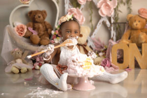 A baby in a white outfit and floral headband sits on the floor, happily eating cake with her hands and a spoon. Surrounding her are teddy bears, pink flowers, and large letters spelling "ONE"—perfect for a cake smash photographer Jacksonville FL.