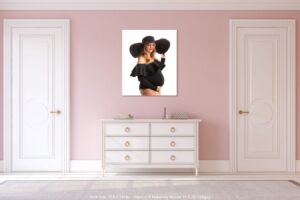 A room with light pink walls, white double doors, and a white dresser. Above the dresser hangs a photo taken by a maternity photographer in Jacksonville FL of a pregnant woman in a black dress and wide-brimmed hat, gently smiling at her belly.