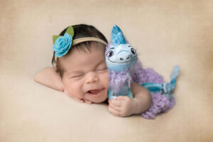 Smiling baby with a blue flower headband lies on a beige surface, holding a blue and purple stuffed dragon toy—captured at a newborn photography studio in Jacksonville, FL—with eyes closed and mouth slightly open in a joyful expression.