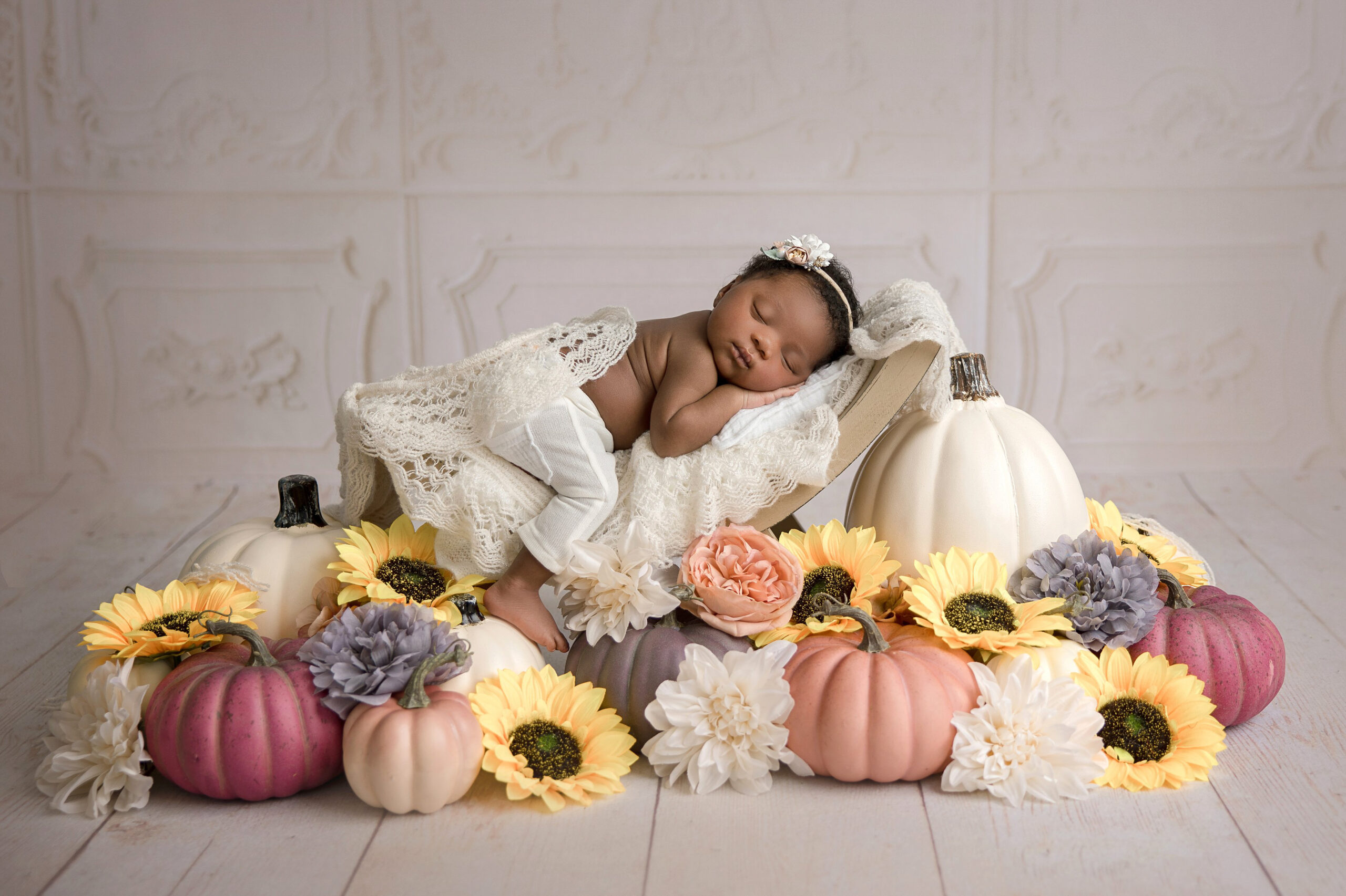 A baby in a lace outfit sleeps on a white pillow, surrounded by pumpkins and sunflowers, creating a soft, elegant scene—perfect inspiration for your newborn photography session in Jacksonville Florida.
