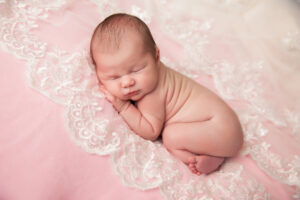 Newborn baby peacefully sleeping on a pink, lace-decorated blanket, curled up on their side—captured beautifully by a Jacksonville baby photographer.