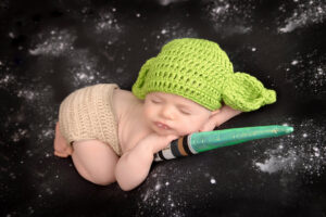 A sleeping baby dressed in a green knit hat with Yoda-like ears and beige shorts lies on a starry black background, cuddling a toy lightsaber—captured by a Jacksonville baby photographer.