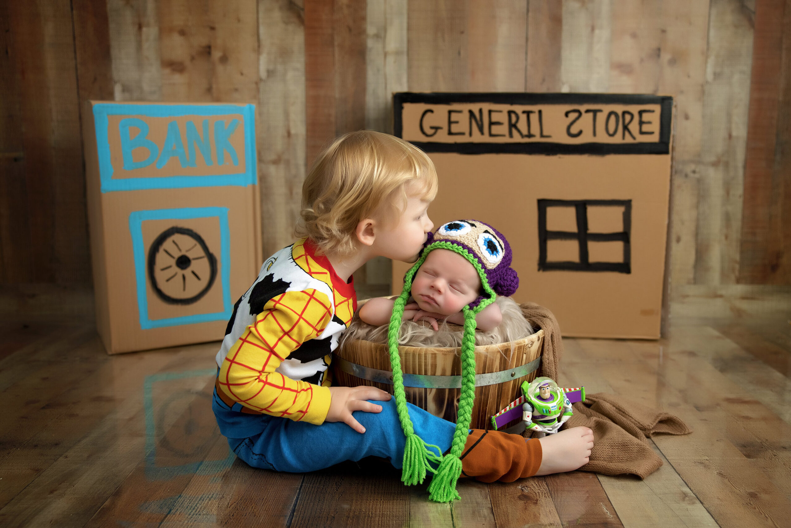 A young child dressed as Woody from Toy Story kisses a baby wearing a colorful hat, who is sleeping in a wooden bucket. Perfect for a newborn photography session in Jacksonville, Florida, with charming props labeled “BANK” and “GENERAL STORE” in the background.